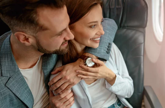 Ein Mann und eine Frau sitzen zusammen in einem Flugzeug. Der Mann umarmt die Frau, beide schauen aus dem Fenster.