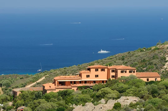 Das rote Gebäude des Hotel Rocce Sarde auf einem Hügel, im Hintergrund das blaue Meer 